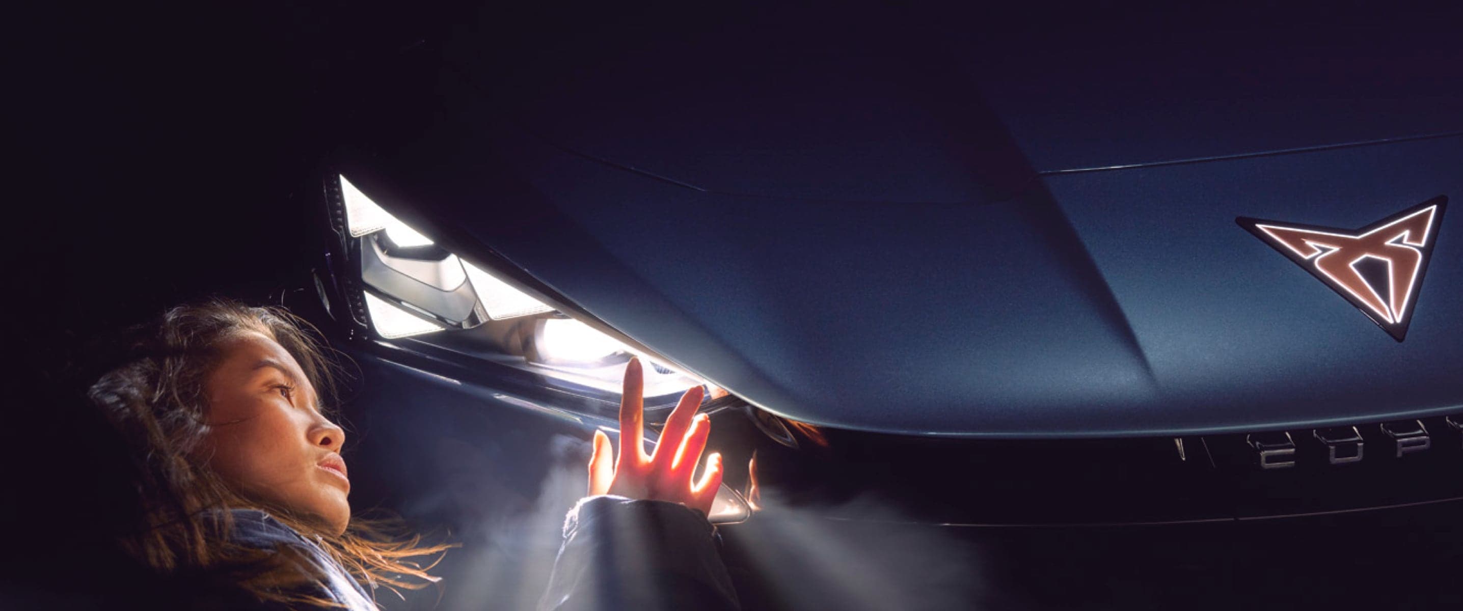 Mujer inspeccionando el interior del capó de un CUPRA Formentor con iluminación y el logotipo CUPRA en primer plano.