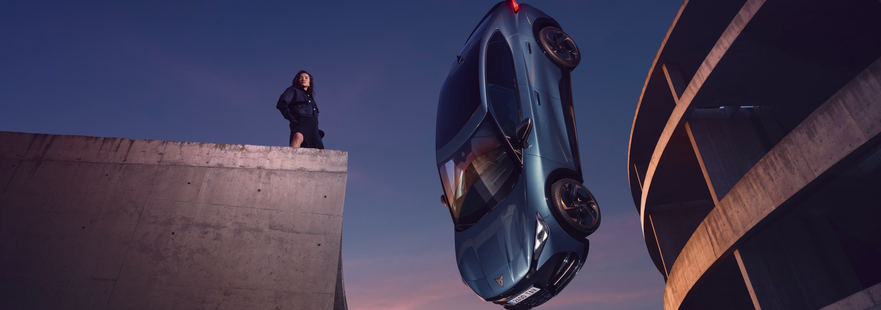 CUPRA Born en azul brillante suspendido en el aire junto a una estructura arquitectónica moderna, con una mujer observando desde un nivel superior.