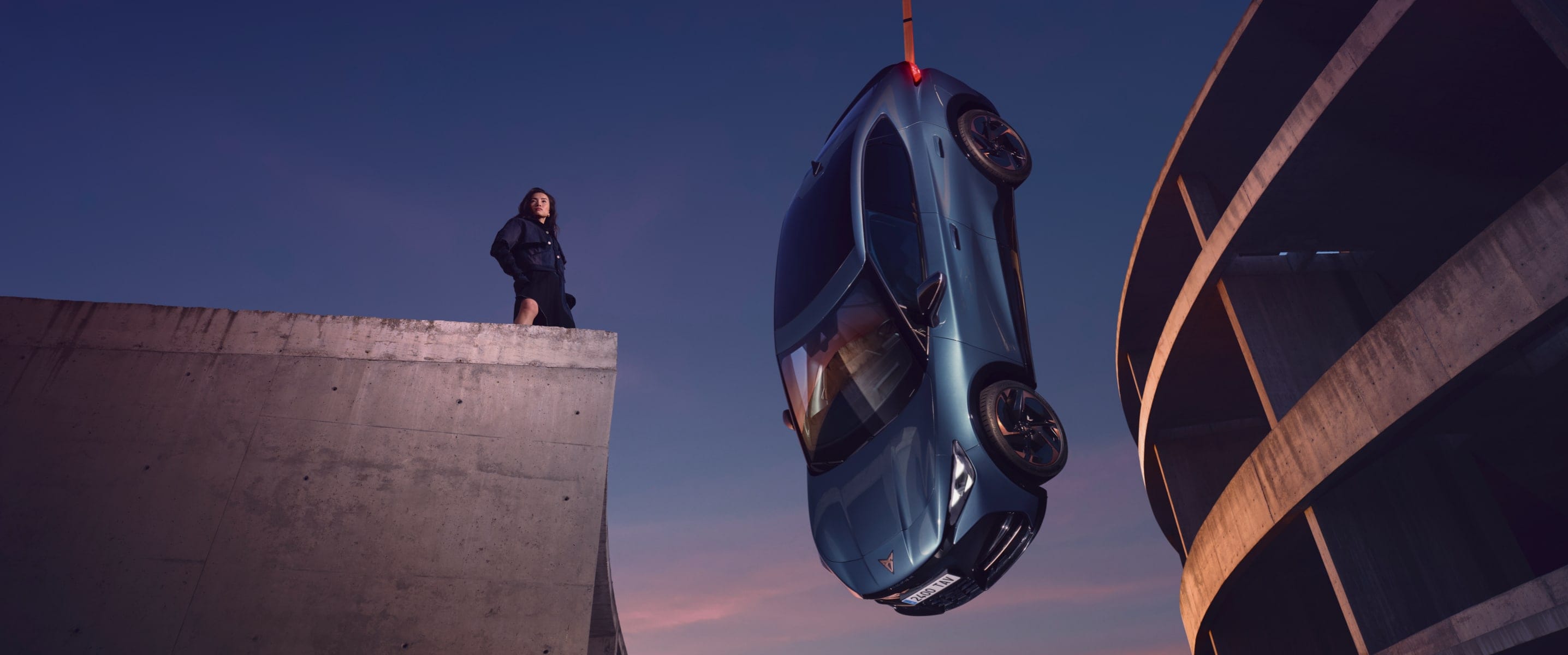 CUPRA Born en azul brillante suspendido en el aire junto a una estructura arquitectónica moderna, con una mujer observando desde un nivel superior.
