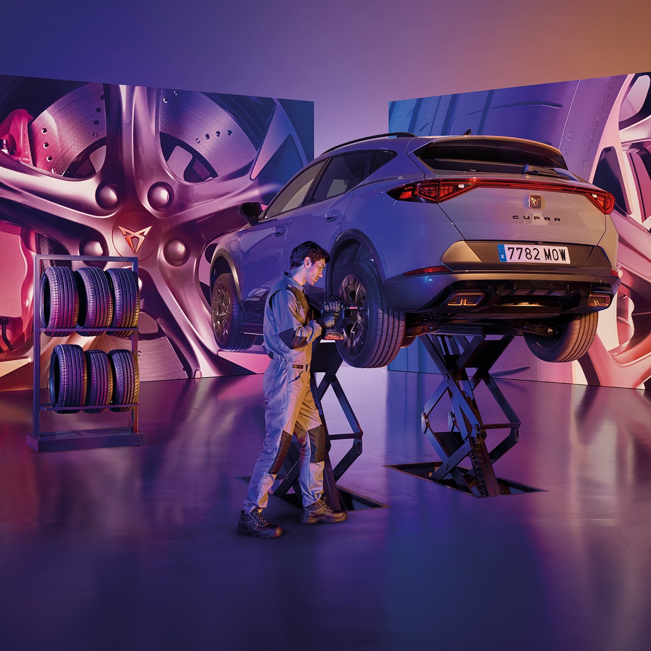 CUPRA Formentor elevado en un taller especializado, rodeado de neumáticos y luces futuristas en púrpura y naranja, mientras un técnico verifica el estado del coche.