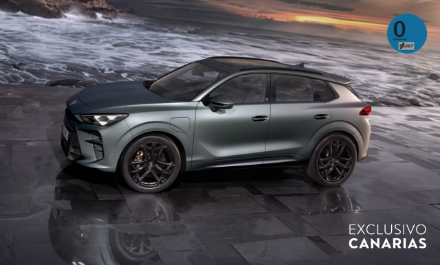 CUPRA Terramar e-HYBRID en color gris estacionado en un entorno urbano con suelo de piedra y fondo costero, destacando su diseño deportivo y tecnología híbrida enchufable.