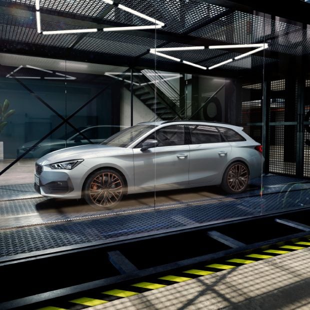 Vista cenital de un coche CUPRA sobre un elevador en un taller de servicios. Una persona está al lado de varias ruedas y una carretilla está cerca del coche al frente. El suelo refleja sutilmente las luces