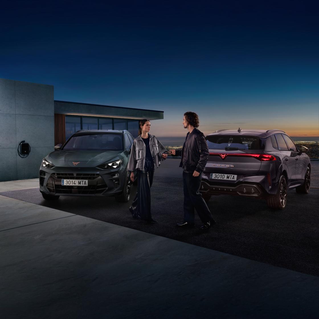 Hombre y mujer están hablando frente a dos CUPRA Terramar híbridos enchufables en un mirador al anochecer