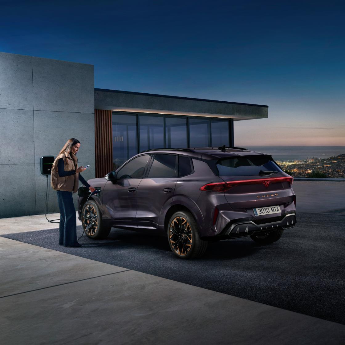 Mujer conectando un CUPRA Terramar híbrido enchufable a punto de carga en casa junto a mirador al anochecer