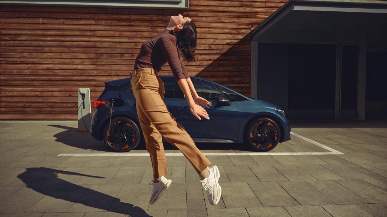 Mujer salta en frente de CUPRA Born 100% eléctrico que está conectado a punto de carga en casa