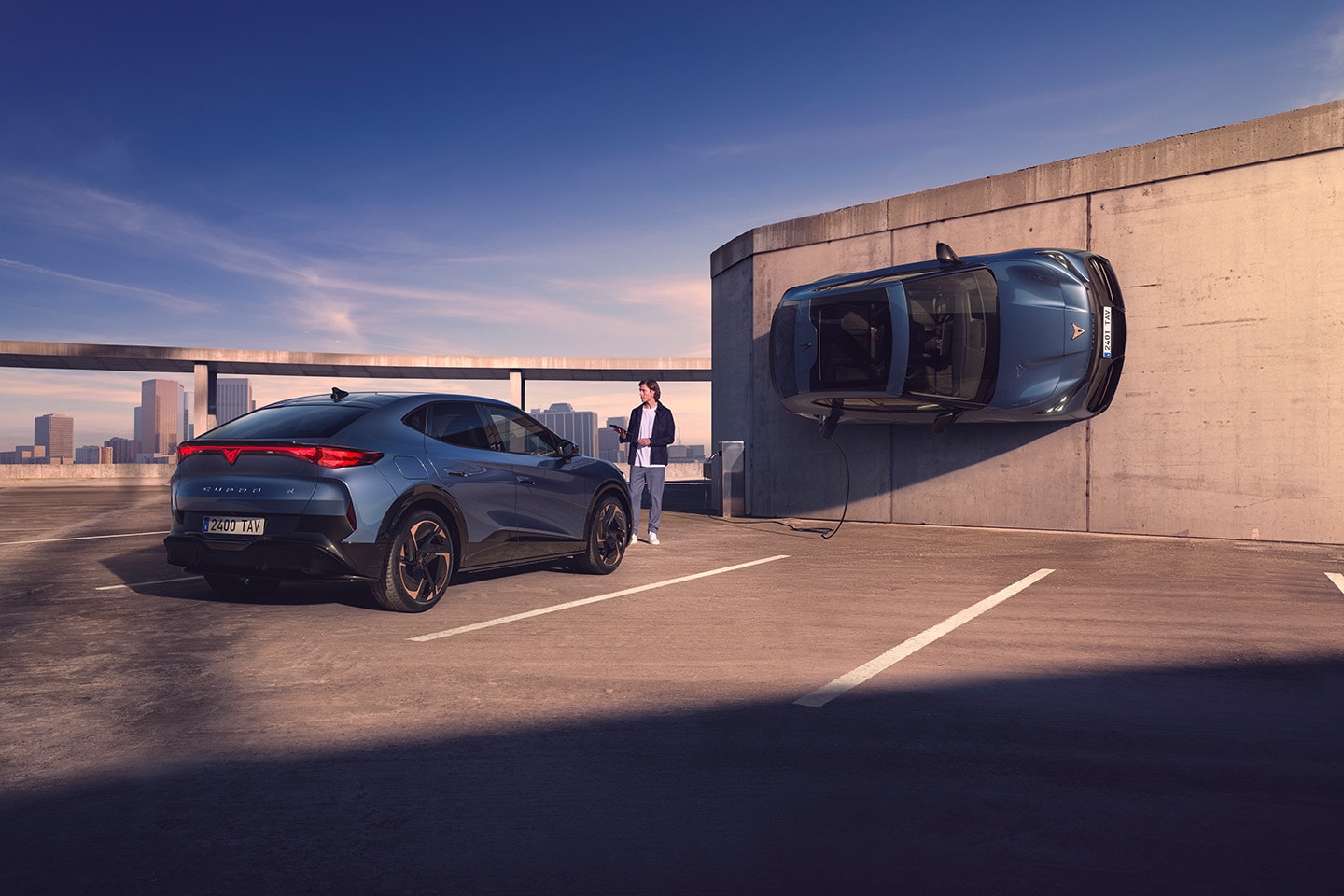 Hombre de pie junto a dos CUPRA Tavascan eléctricos, uno conectado a cargador público mientras está colgado en la pared y el otro aparcado en plaza aparcamiento
