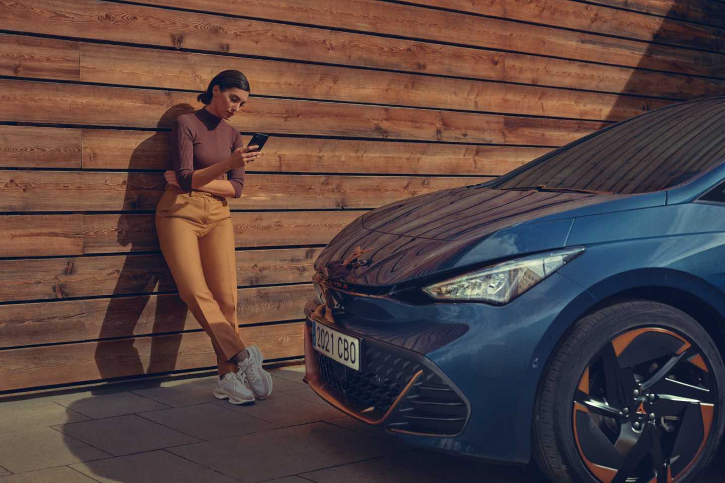 Mujer navega en su smartphone de pie junto a un CUPRA Born eléctrico de color azul