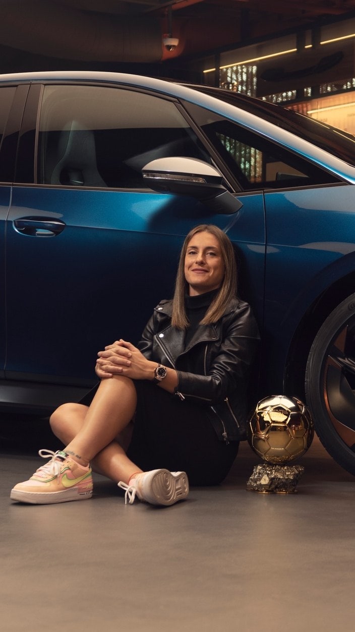 Alexia Putellas sentada en el suelo junto trofeo Balón d’Or apoyada en el lateral de un CUPRA Born
