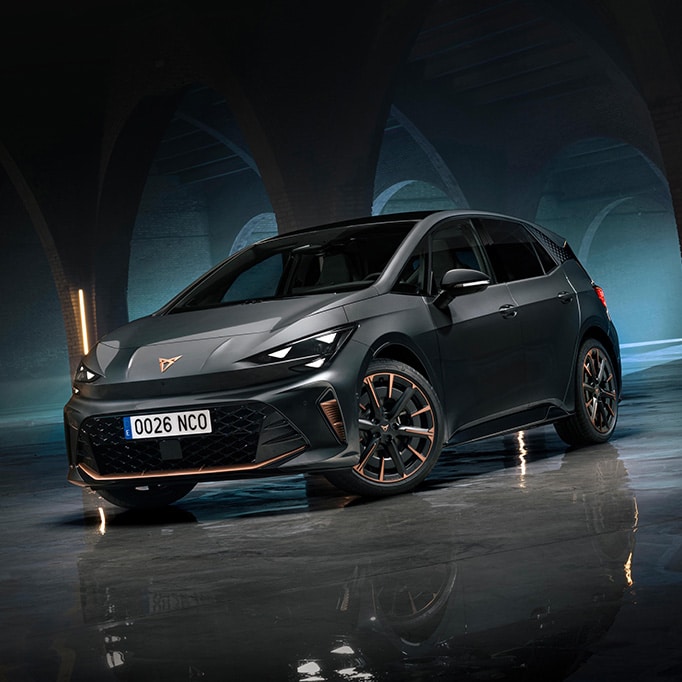    A front-three-quarter view of a CUPRA Born VZ in Timanfaya Grey with copper accents on the wheels and grill, parked in a dimly lit industrial space featuring large stone arches and vertical neon light bars.