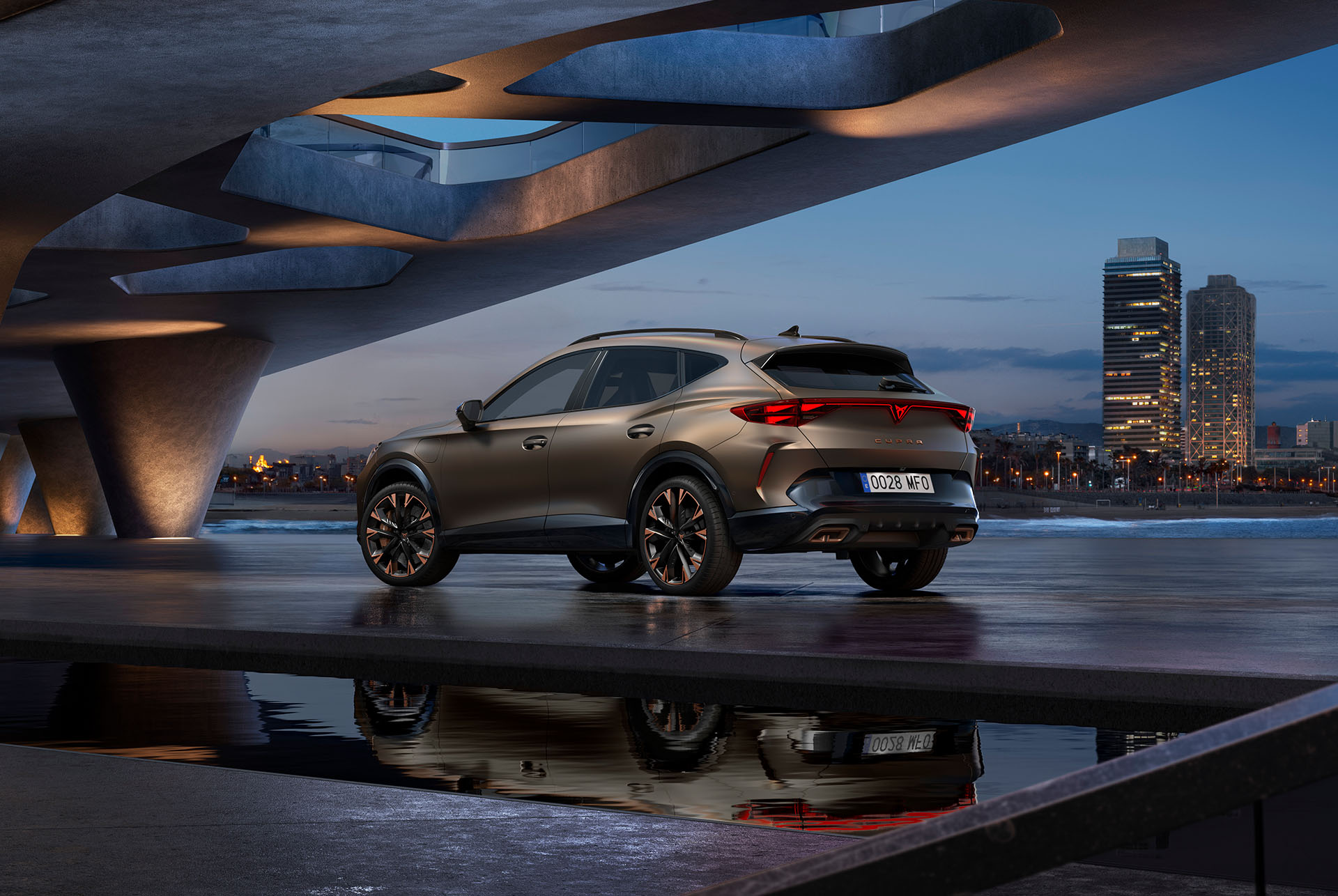 Perspectiva trasera de tres cuartos del nuevo coche híbrido CUPRA Formentor aparcado junto al paseo marítimo y el skyline de la ciudad. El vehículo está inclinado para que se vea su diseño trasero, las luces y los tubos de escape dobles deportivos.