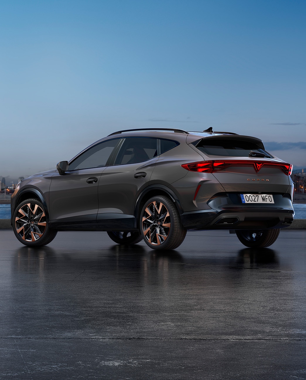 El nuevo SUV crossover CUPRA Formentor 2025 gris Graphene aparcado en un paseo marítimo durante el atardecer. El fondo muestra un paisaje urbano con rascacielos y palmeras iluminados por la luz del día, que se va atenuando.