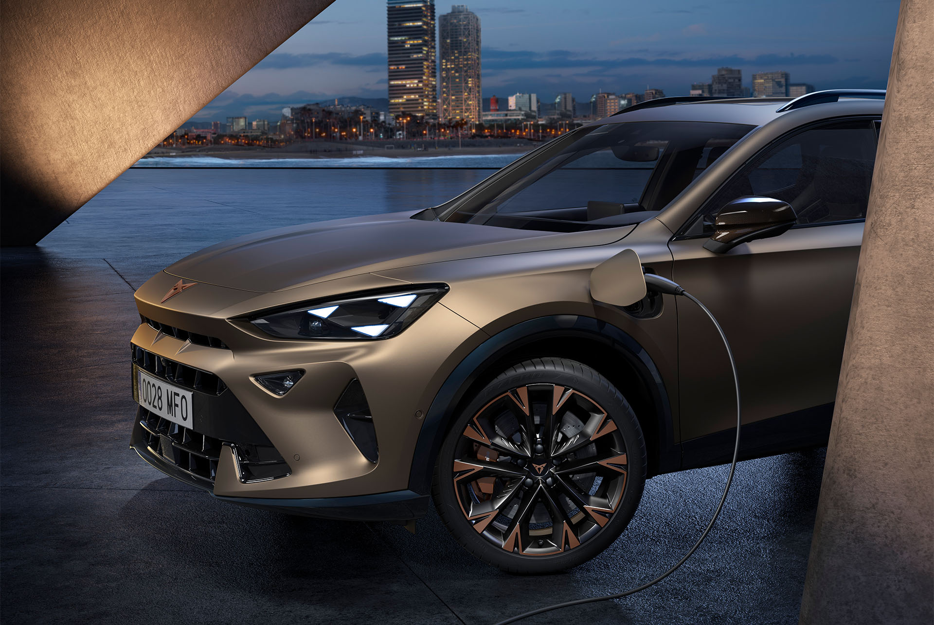 Vista frontal izquierda del nuevo CUPRA Formentor 2025 Century Bronze mate con llantas en acabado Copper, cargándose, con una vista de fondo del paisaje urbano de Barcelona al atardecer.