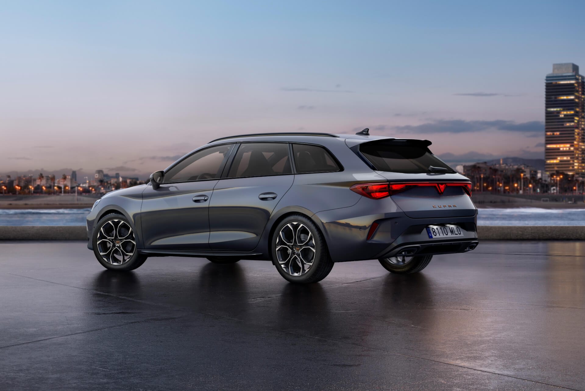 Vista trasera del nuevo modelo de coche CUPRA León Sportstourer 2025 gris brillante con control de chasis adaptativo y llantas de aleación en negro y cobre. Ciudad al fondo al atardecer.