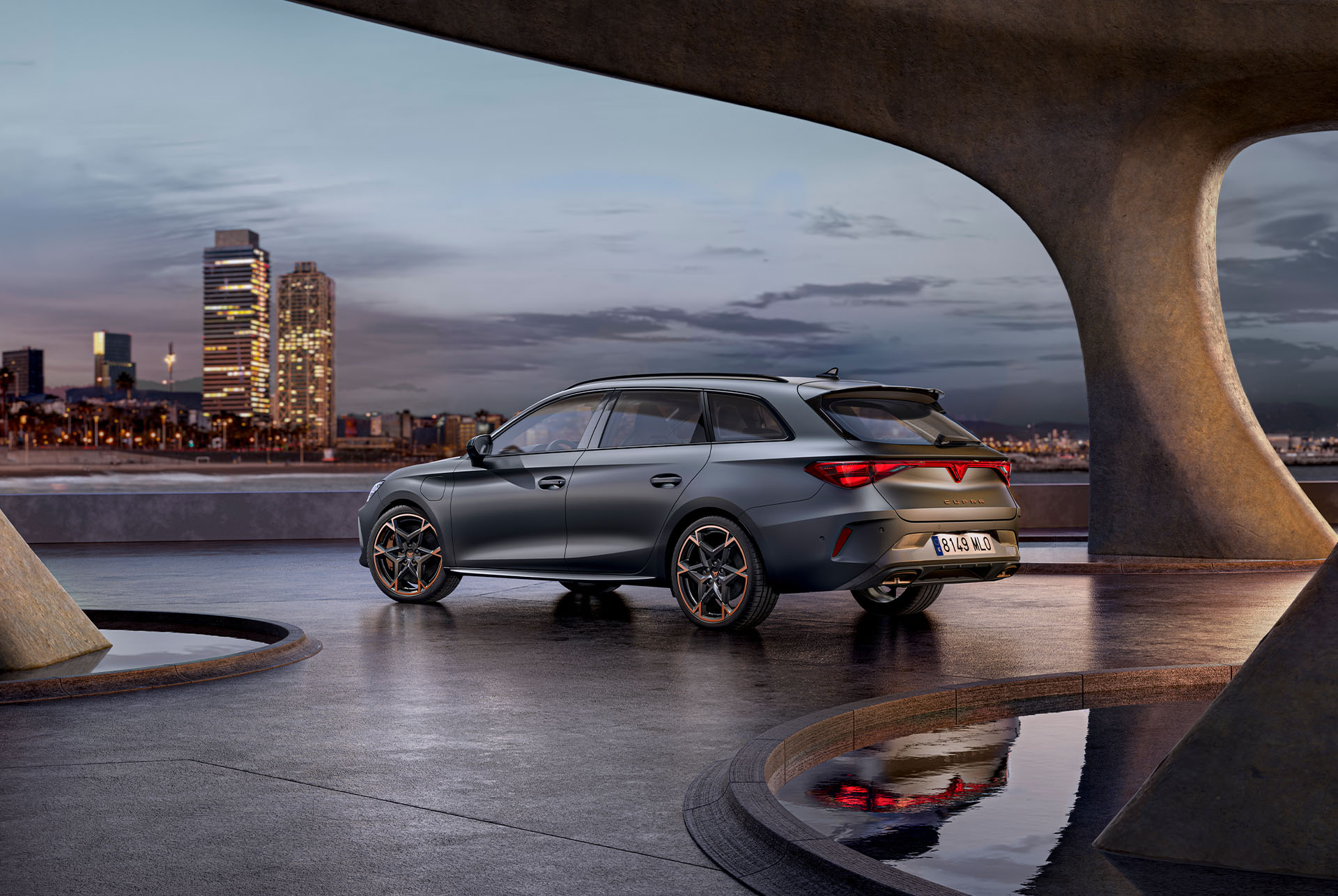 Vista lateral trasera del nuevo vehículo deportivo y aerodinámico CUPRA León Sportstourer 2025 gris mate, aparcado en un paseo marítimo al atardecer. El skyline de la ciudad reluce al fondo.
