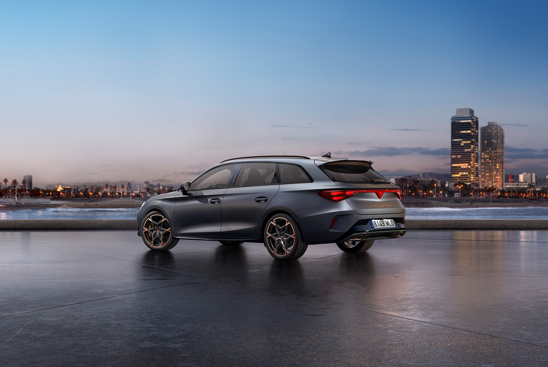 The sporty Cupra Leon Sportstourer parked on concrete with a city skyline illuminated at twilight in the background. The vehicle is depicted in a matte grey finish.