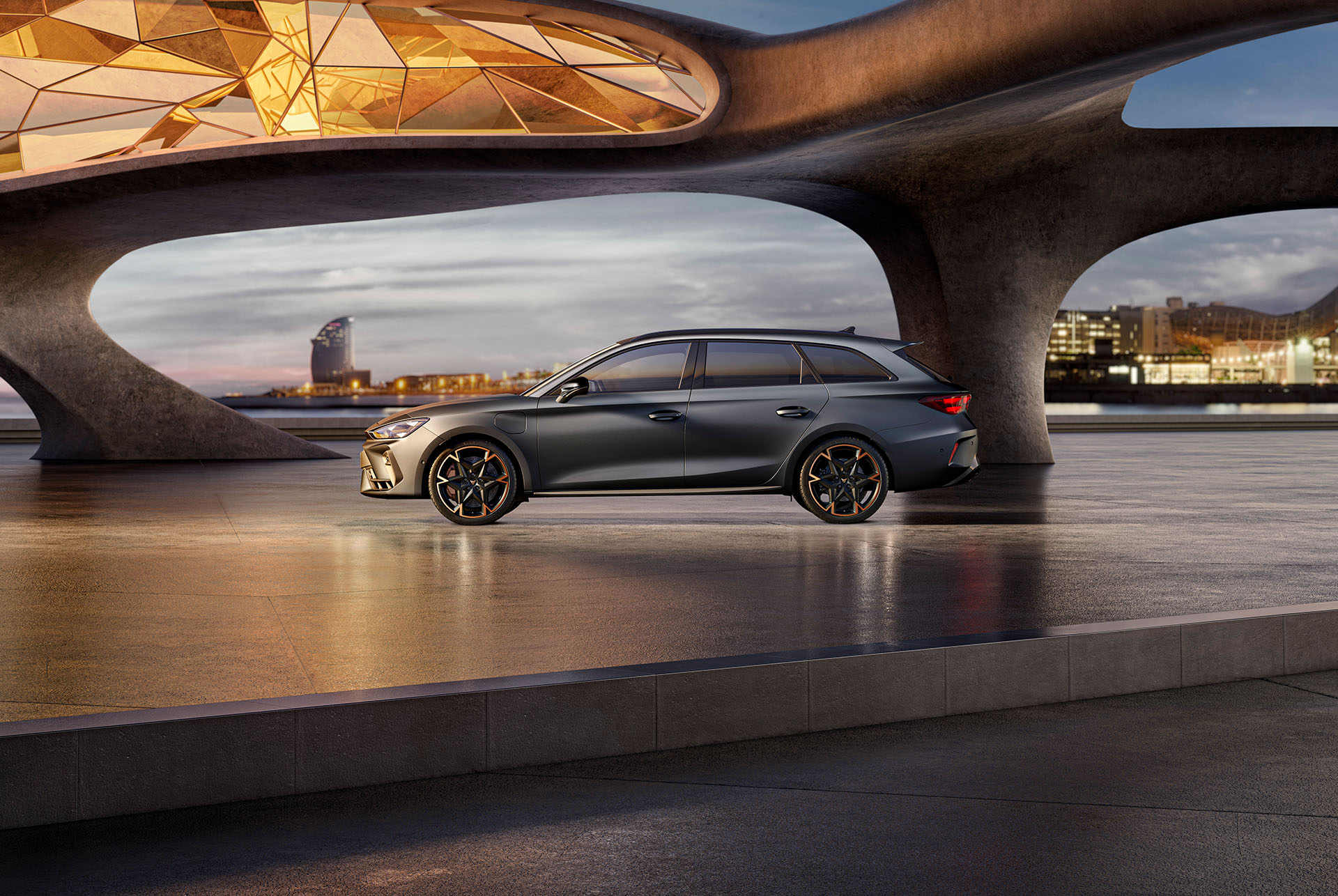 Nuevo CUPRA León Sportstourer 2025, versiones y equipamiento, aparcado sobre cemento, estructura con bordes brillantes, con vistas al skyline de una ciudad al atardecer. El vehículo se muestra en un acabado gris mate con llantas de aleación de color cobre y negro.
