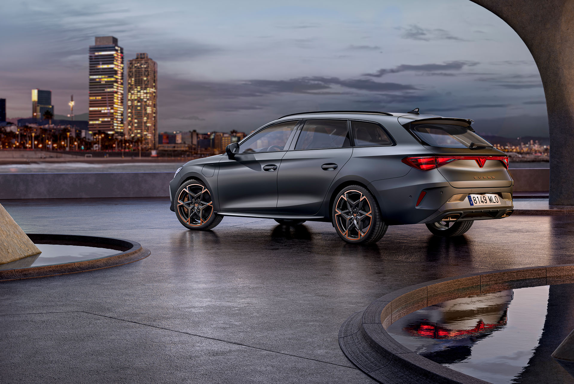 Tecnología del nuevo CUPRA León Sportstourer 2025 estacionado sobre cemento, estructura con bordes brillantes, con vistas al skyline de una ciudad al atardecer. El vehículo se muestra en un acabado gris mate con llantas de aleación de color cobre y negro.