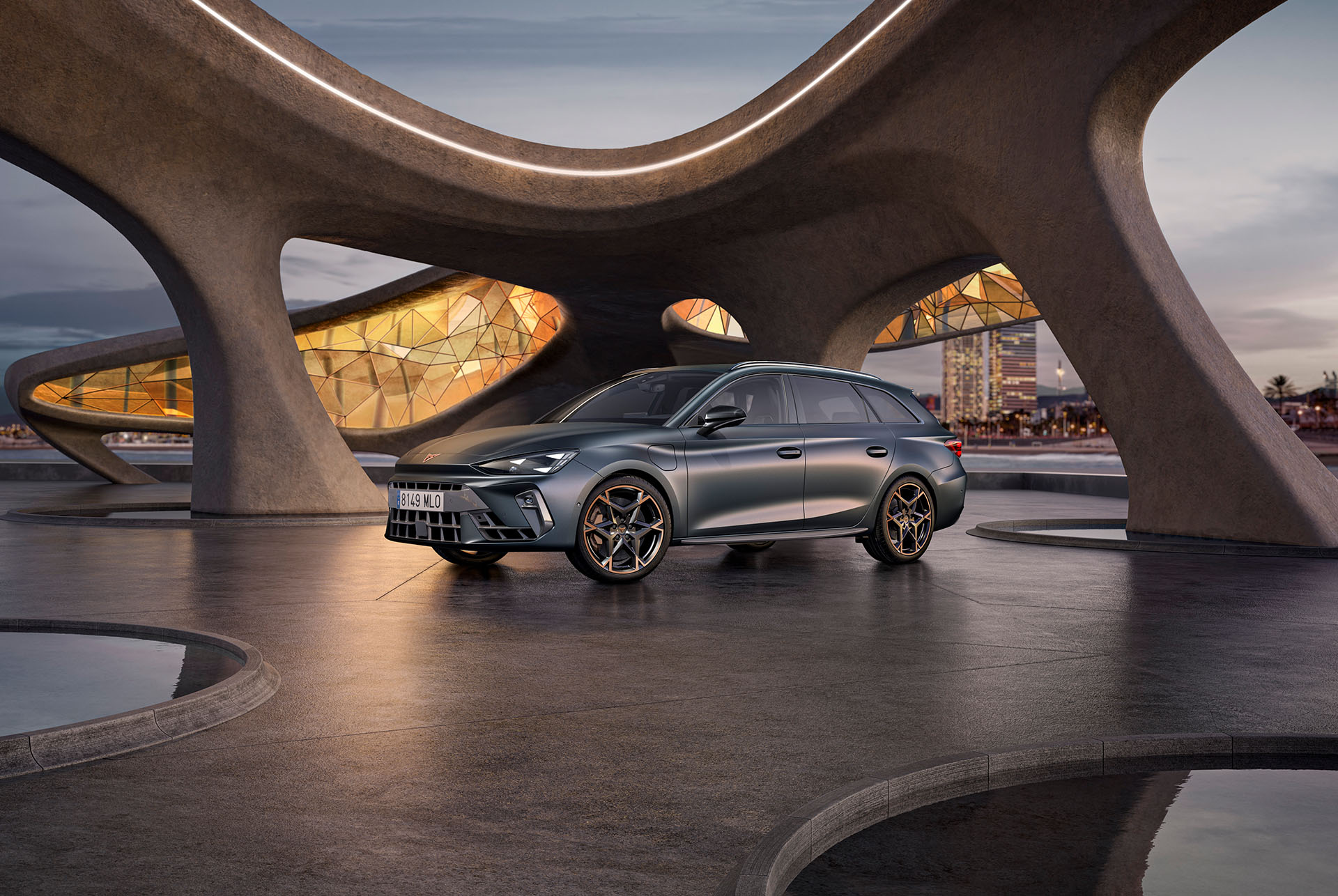 Vista gran angular del nuevo y deportivo vehículo híbrido CUPRA León Sportstourer 2025 en gris mate, aparcado en un entorno urbano, delante de grandes ventanales geométricos inspirados en Gaudí.