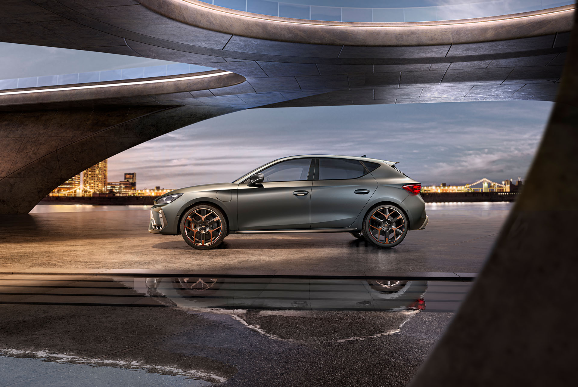 Plano general del nuevo CUPRA León 2025, en gris, vista lateral, aparcado sobre hormigón liso bajo una estructura arquitectónica con vistas al mar y paisaje urbano, cielo nublado.