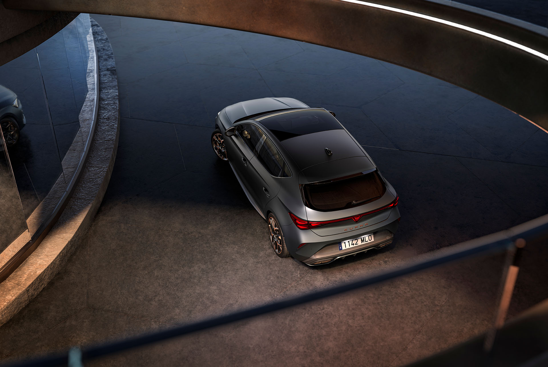 Tecnología del nuevo CUPRA León e-Hybrid 2025, iluminación trasera LED, vehículo Híbrido Enchufable aparcado sobre cemento, estructura con bordes brillantes. El vehículo se muestra en acabado gris mate con llantas forjadas en cobre y negro.