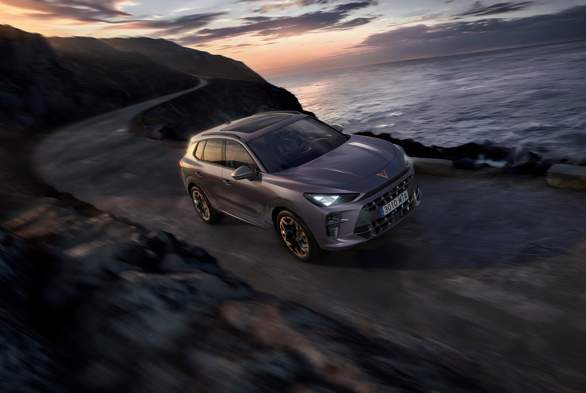 CUPRA Terramar color Dark Void con llantas de aleación Hadron Copper de 20 pulgadas, faros CUPRA Matrix LED ultra HD y Control de chasis adaptativo (DCC), conduciendo por una carretera sinuosa mojada junto al mar al atardecer.