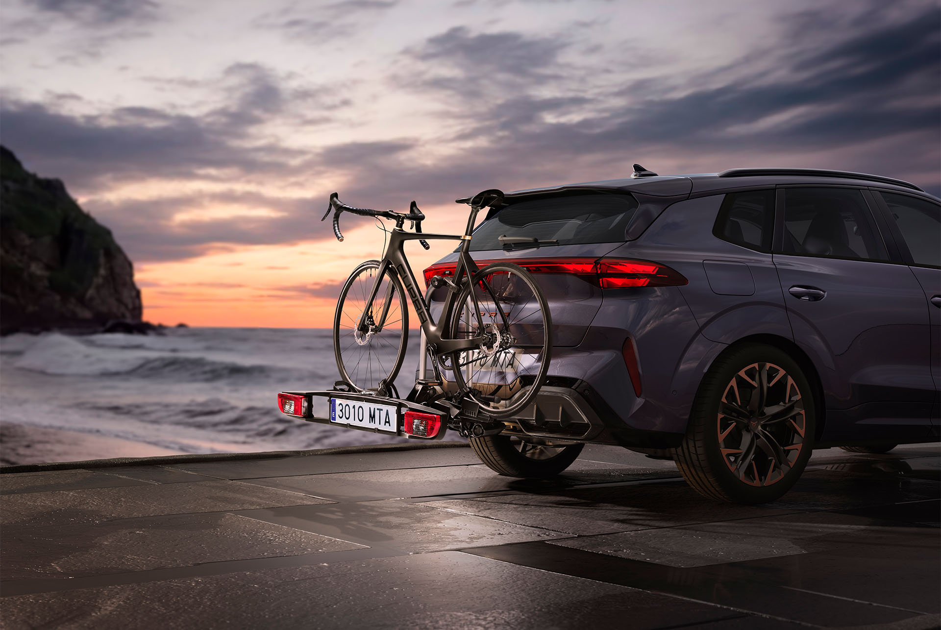 Remolque portabicicletas CUPRA con dos bicicletas enganchadas, aparcado junto a la costa al atardecer. El remolque portabicicletas ofrece una opción cómoda y segura para transportar bicicletas, capaz de soportar hasta 60 kg. 
