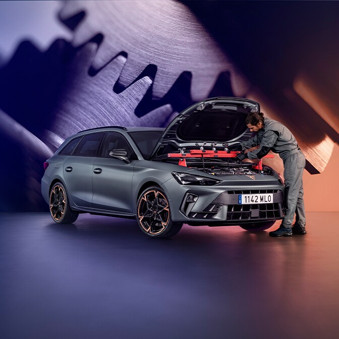 Un técnico vestido con un mono gris inspecciona el compartimento del motor de un CUPRA Terramar en un taller; al fondo se ve una gran pantalla en la que se muestra un primer plano de unos engranajes metálicos.