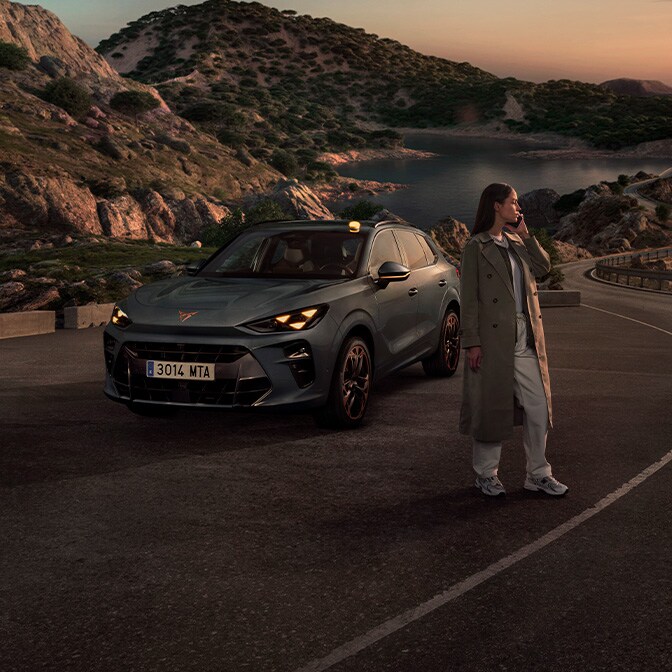 Una fotografía panorámica tomada al atardecer en la que se ve a una mujer con gabardina de pie en una sinuosa carretera costera junto a un CUPRA Terramar, con colinas rocosas y un mar en calma al fondo.