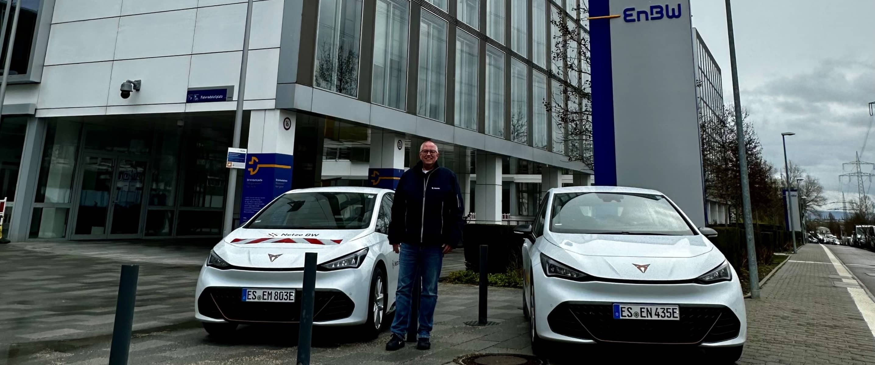 Dos vehículos CUPRA Born blancos aparcados en el exterior de un edificio de oficinas de EnBW, con un administrador de flotas de pie entre los coches, en un día nublado.