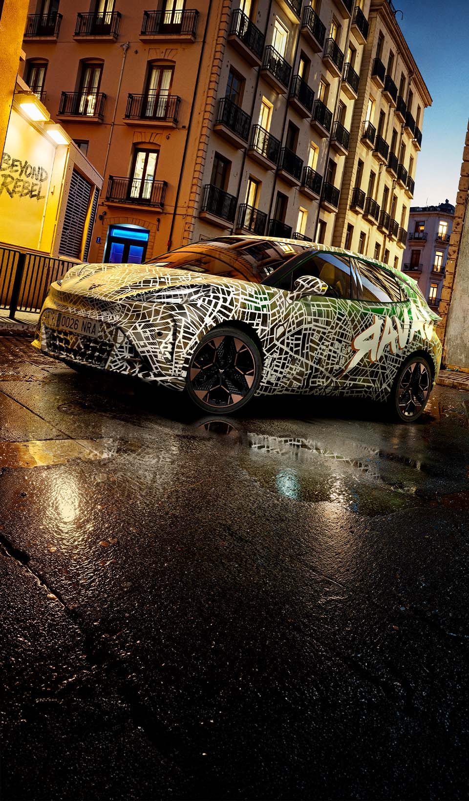 Un prototipo CUPRA Raval se encuentra en una calle urbana mojada y poco iluminada, envuelto en un patrón de camuflaje que recuerda a un mapa callejero blanco y verde.