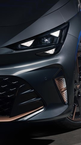 A detailed close-up of the front left headlight and bumper of a CUPRA Born VZ in Timanfaya Grey, showcasing the sharp, triangular LED daytime running lights and copper-finished accents on the lower grille and alloy wheel.