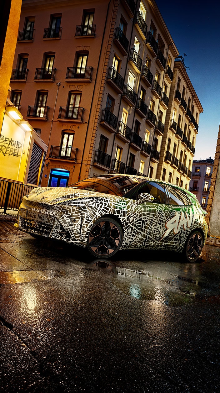 CUPRA Raval con vinilo de camuflaje, que combina patrones abstractos y la palabra “Raval”, estacionado en una calle mojada al anochecer, con altos edificios residenciales iluminados al fondo.