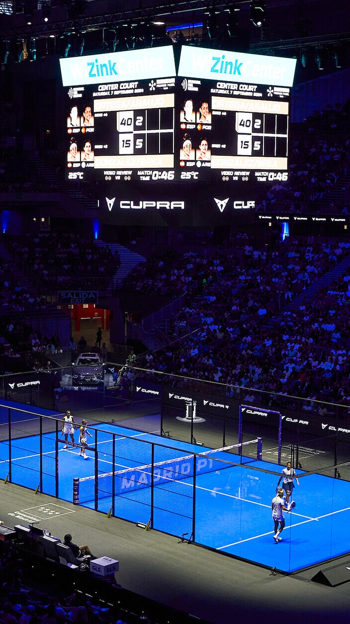 Una pista de pádel iluminada por luces azules, vista desde arriba durante un partido dentro de un gran estadio lleno de espectadores. Dos parejas de jugadores se encuentran en la pista con un gran marcador en el fondo