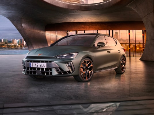 CUPRA León e-HYBRID 5 puertas color gris estacionado frente a una arquitectura contemporánea con iluminación dorada.