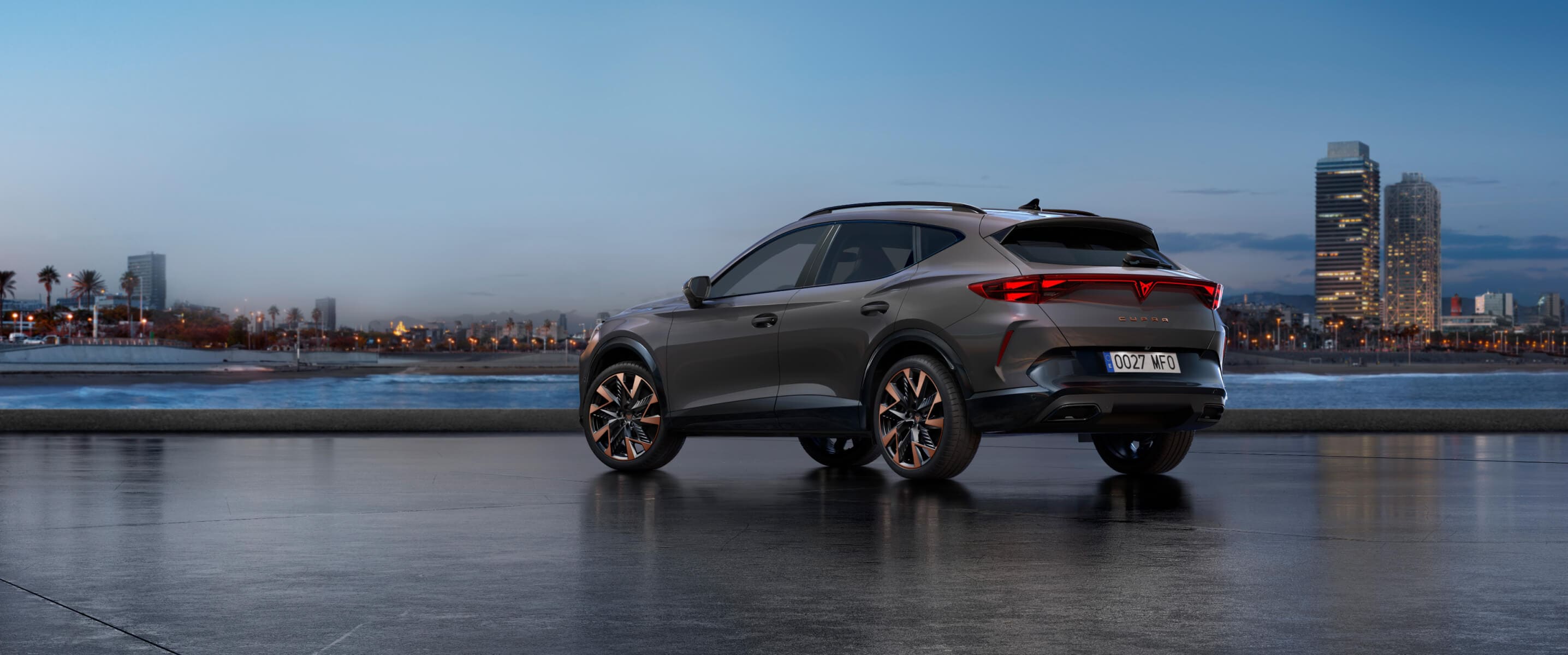 Qué es un coche microhíbrido, CUPRA Formentor en gris Graphene, vista trasera aparcado junto al agua al atardecer con el skyline de la ciudad.
