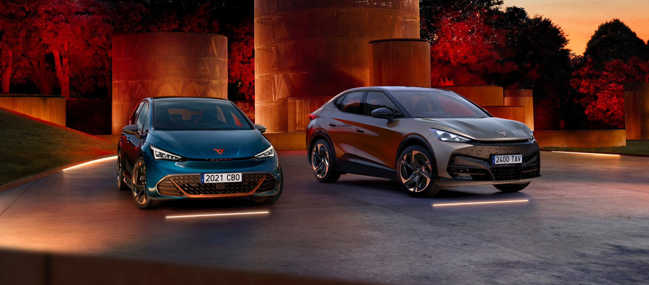 Un CUPRA Born y un CUPPRA Tavascan, aparcados al aire libre en un parque, rodeados de luces tenues.